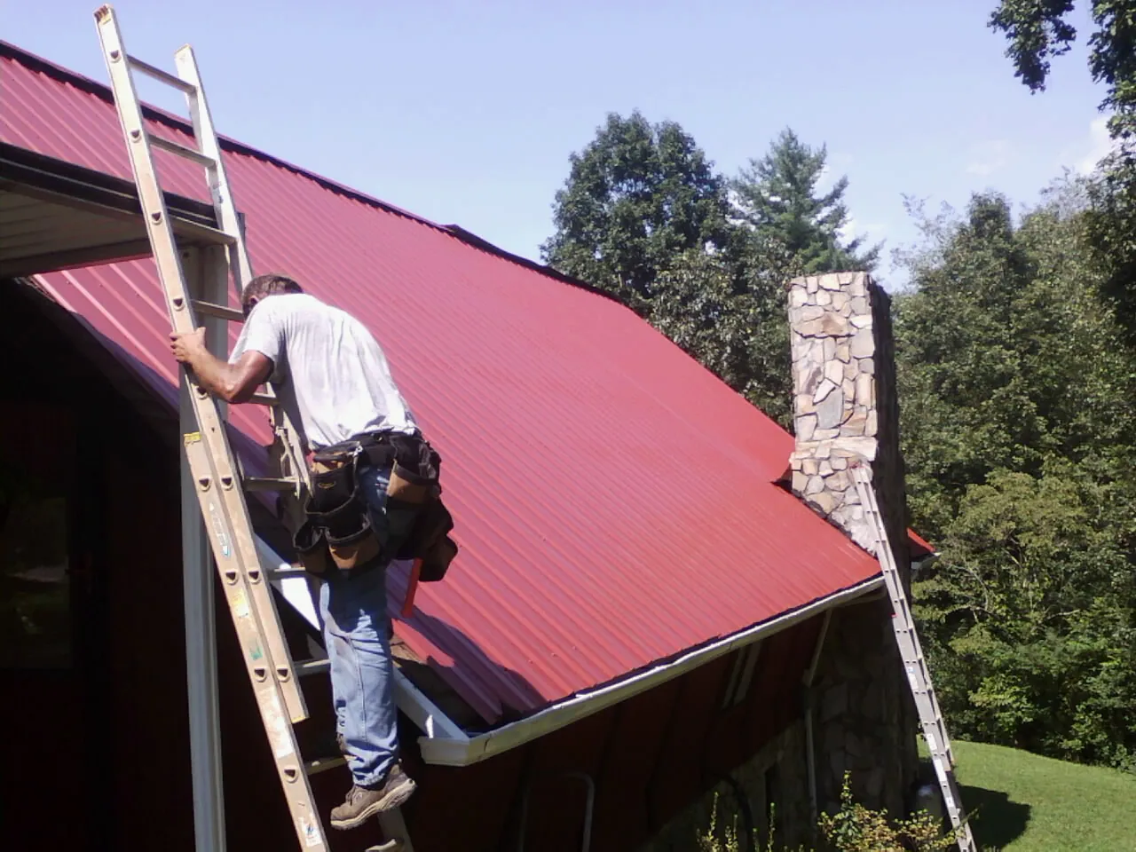 Roofer on a ladder beside a red metal roof and stone chimney — Soffit & Fascia Repair by Roofing Brothers in Verdon